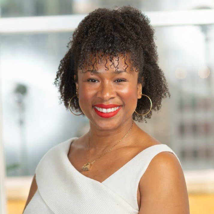 Woman with curly hair and hoop earrings smiles at the camera, wearing a white sleeveless top, standing indoors with a bright, blurred background.